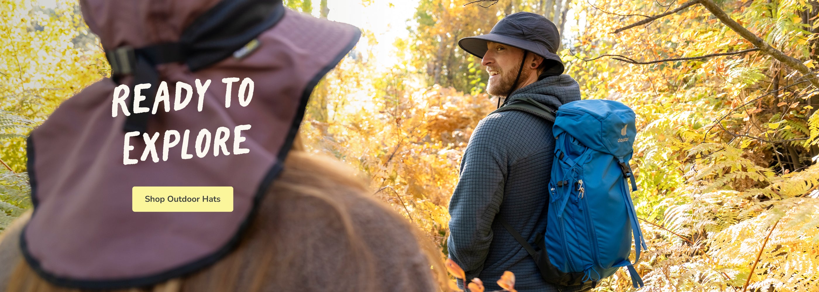 A man wearing a dark wide-brim hat and a blue hiking backpack walks through a sunlit autumn forest, while another person in the foreground wears a maroon sun hat with the text 'READY TO EXPLORE' and a 'Shop Outdoor Hats' button.